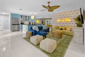 A modern living room with a blue sofa, yellow sofa, and a green rug. There is a kitchen area with a refrigerator and a ceiling fan. - Private Pool Movie Theater Putting Playground (Seminole)