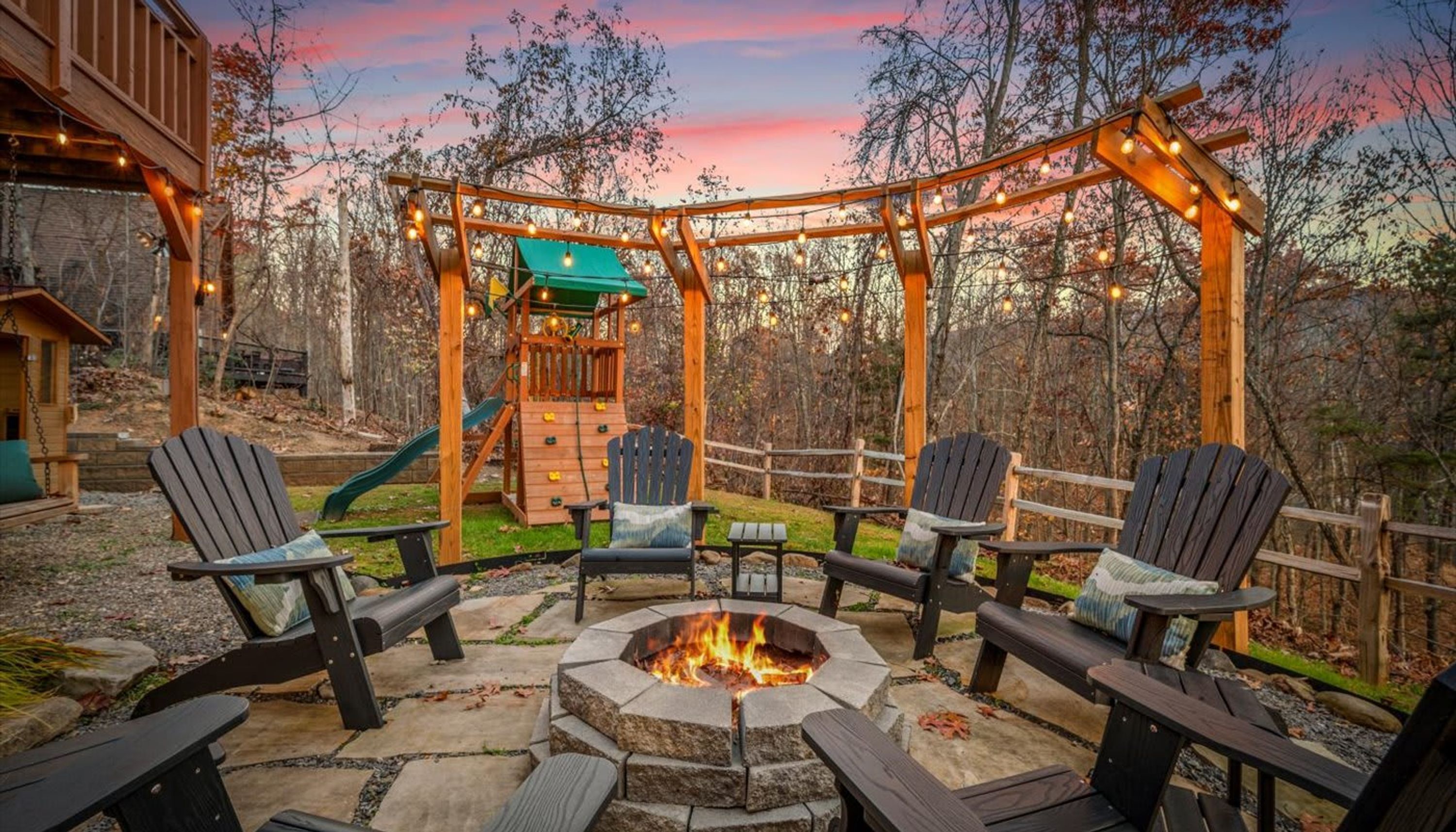 Un espacio exterior con sillas Adirondack, un brasero y una pérgola de madera con luces colgantes.