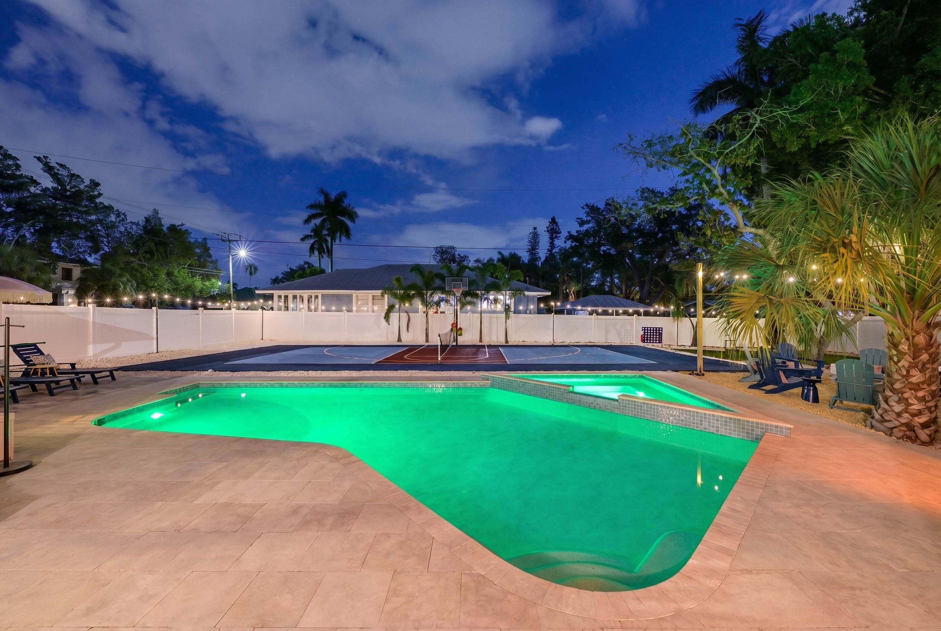 A swimming pool with a curved edge, surrounded by a white fence, and a basketball court in the background.