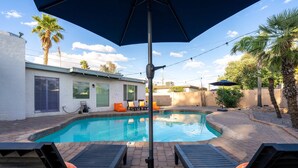 Ein Swimmingpool mit Liegestühlen und Sonnenschirm, umgeben von Palmen und einem Gebäude.