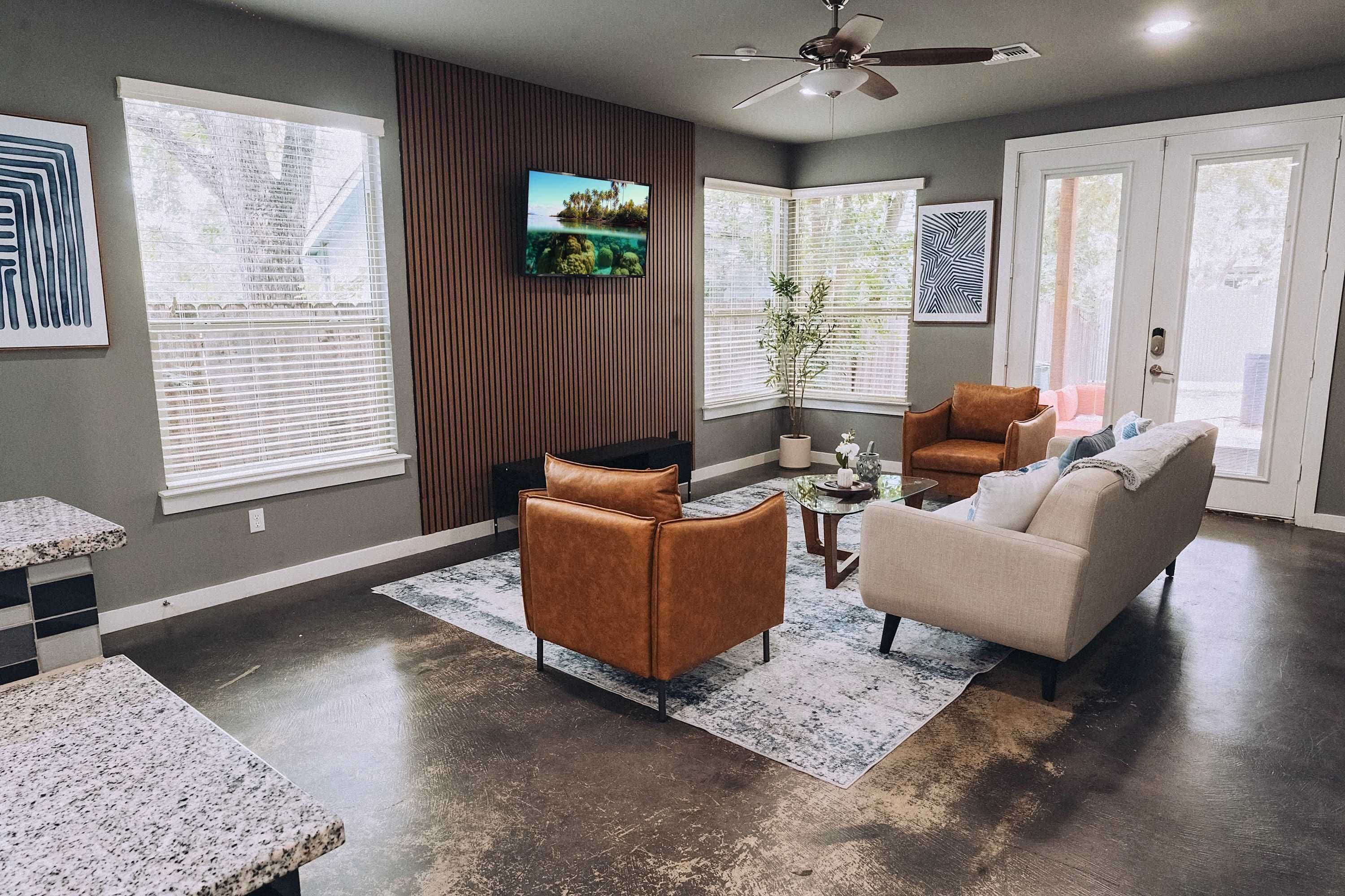 A modern living room with a flat-screen TV mounted on the wall, a ceiling fan, and a large window with blinds.