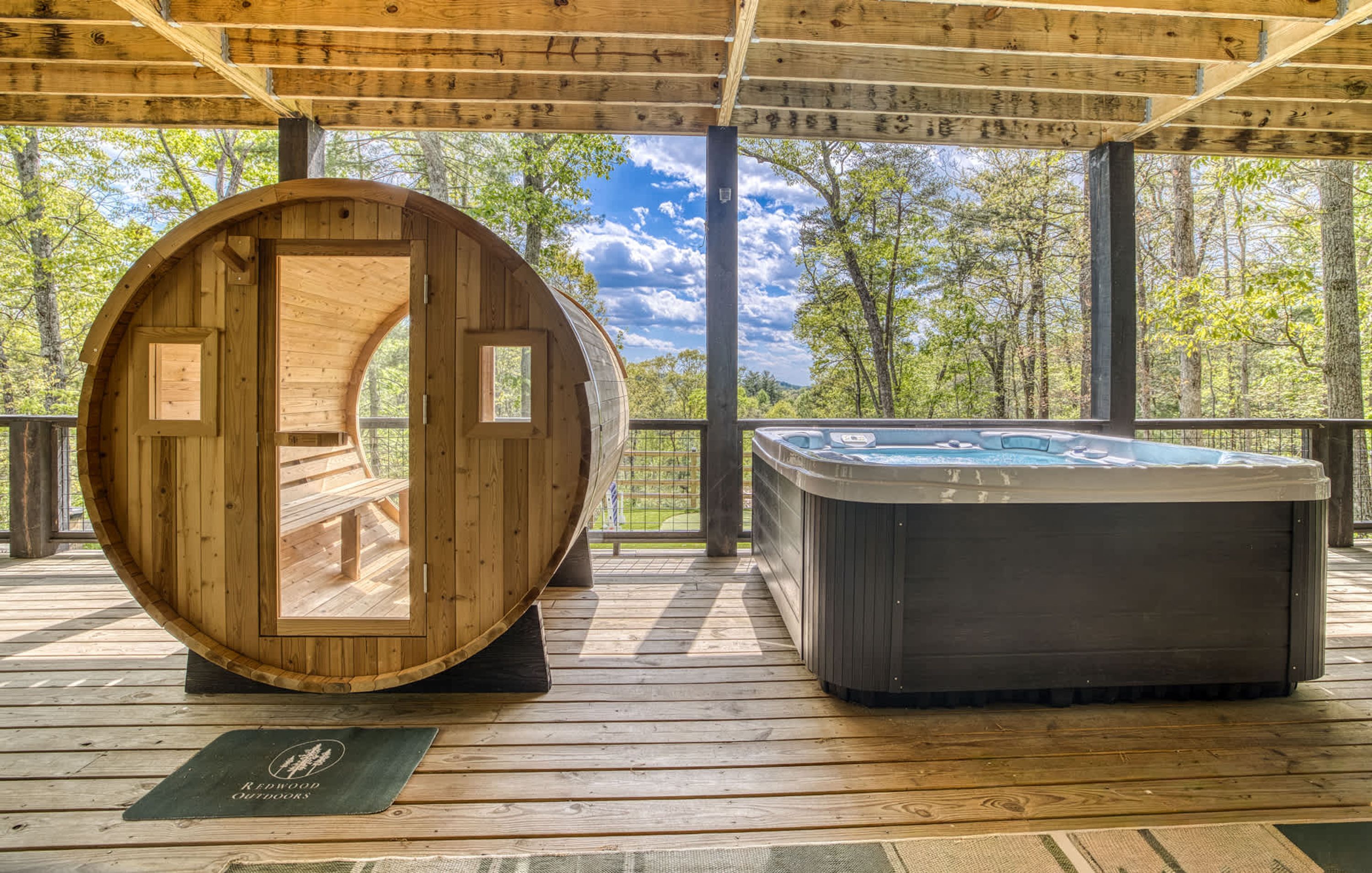 A wooden barrel structure and a hot tub on a wooden deck with a view of trees and sky.