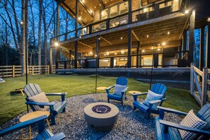 A modern outdoor seating area with Adirondack chairs and a fire pit, surrounded by a wooden deck and a view of a large building with multiple windows. - The Glass Cabin Hot Tub Sauna (Morganton)