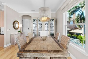 Ein Esszimmer mit Holztisch, Korbstühlen, einer Chandelüster und Blick auf einen Garten durch ein großes Fenster.