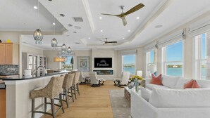 Un salón espacioso con un sofá blanco grande, una zona de comedor con taburetes de bar, una cocina con isla central y un televisor montado en la pared.