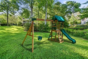 Ein Spielplatz mit Schaukel und Rutsche, umgeben von Gras und Bäumen.
