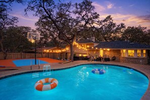 Una piscina junto a una cancha de baloncesto, árboles y una casa con iluminación exterior.