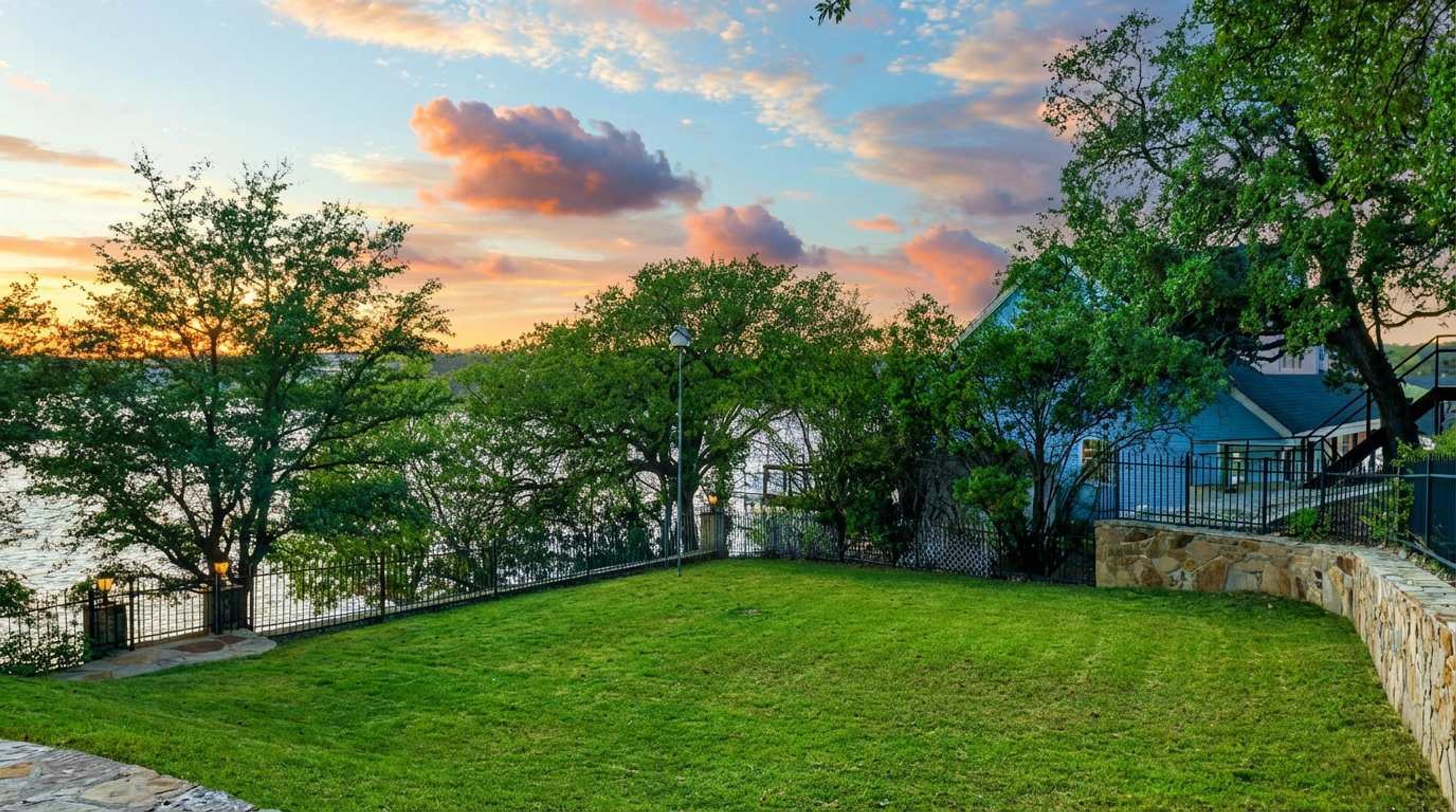 A well-maintained grassy yard with a stone wall and a metal fence, overlooking a body of water and a building with a blue roof.
