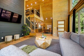 Un vaste espace de vie avec un escalier menant à un niveau supérieur, une grande fenêtre donnant sur la verdure et un coin salon confortable avec une table basse.