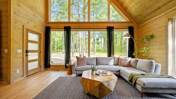 A modern log cabin interior with a large sectional sofa, a coffee table, and a view of a forest through large windows.