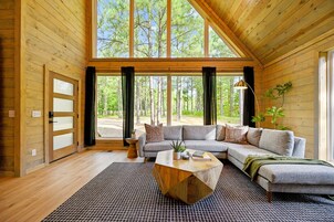 Un interior de cabaña de troncos moderno con un amplio sofá modular, una mesa de centro y vistas a un bosque a través de amplios ventanales.