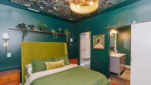 A room with a green bed, a nightstand, a wall-mounted shelf with plants, a door leading to a bathroom, and a ceiling light fixture. - Velvet Haus Downtown 3rd Floor Deck Backyard (Cincinnati)