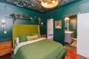 Ein Zimmer mit einem grünen Bett, einem Nachttisch, einem an der Wand befestigten Regal mit Pflanzen, einer Tür zu einem Badezimmer und einer Deckenleuchte.