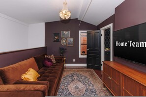 A living room with a brown sofa, a large flat-screen TV, a chandelier, and a door with a sign reading 'Home Team'. - Velvet Haus Downtown 3rd Floor Deck Backyard (Cincinnati)