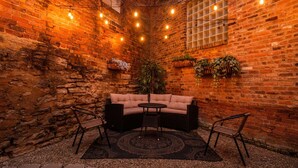 A cozy indoor seating area with a brick wall, a cushioned sofa, two chairs, a small table, and string lights. - Velvet Haus Downtown 3rd Floor Deck Backyard (Cincinnati)