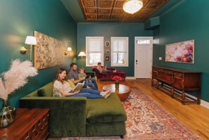 Ein Wohnzimmer mit einem grünen Sofa, einem Holztisch und einem gemusterten Teppich. Zwei Fenster mit Jalousien, eine Tür und eine Deckenleuchte sind ebenfalls zu sehen.