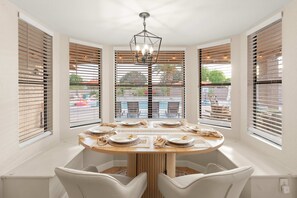Un coin repas avec une table ronde dressée pour quatre personnes, entourée de chaises blanches, avec de grandes fenêtres et des stores sur les côtés.