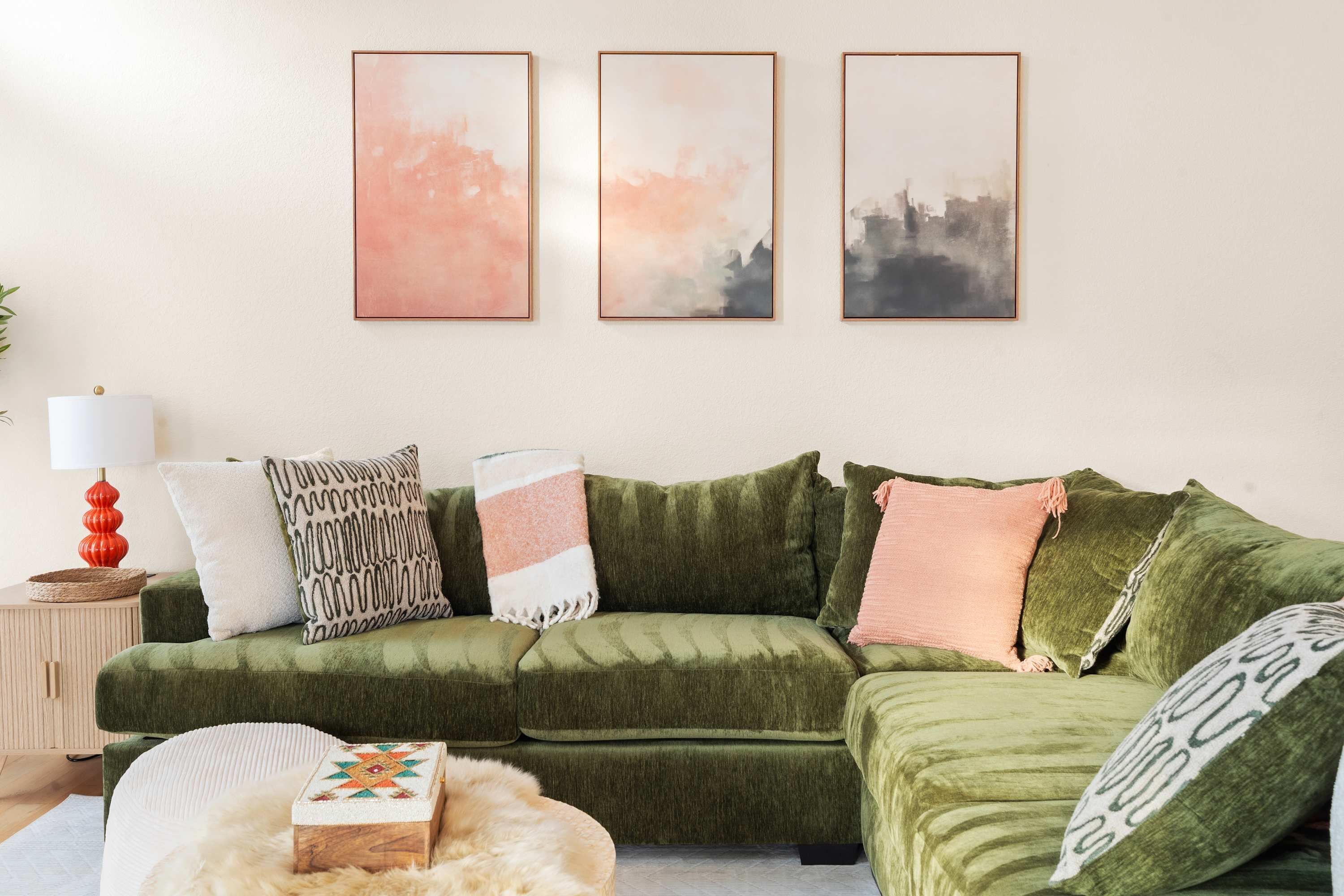 Um sofá verde com várias almofadas, uma pequena mesa redonda com abajur e três quadros abstratos na parede.