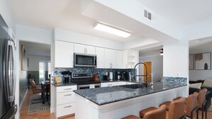 A modern kitchen with white cabinetry, a central island with bar stools, a built-in microwave, and a stainless steel refrigerator. - Texas Tranquility Hot Tub Pool Putt Putt More (San Antonio)