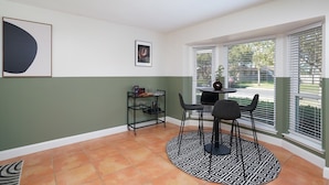 A room with a dining table, chairs, a small shelf with glasses, a framed picture, and a window with blinds. - Texas Tranquility Hot Tub Pool Putt Putt More (San Antonio)