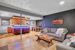 A modern living room with a grey sofa, a wooden coffee table, and a bar area with stools. - Poconos Retreat Hot Tub Firepits Sleeps 16 (Lake Harmony)
