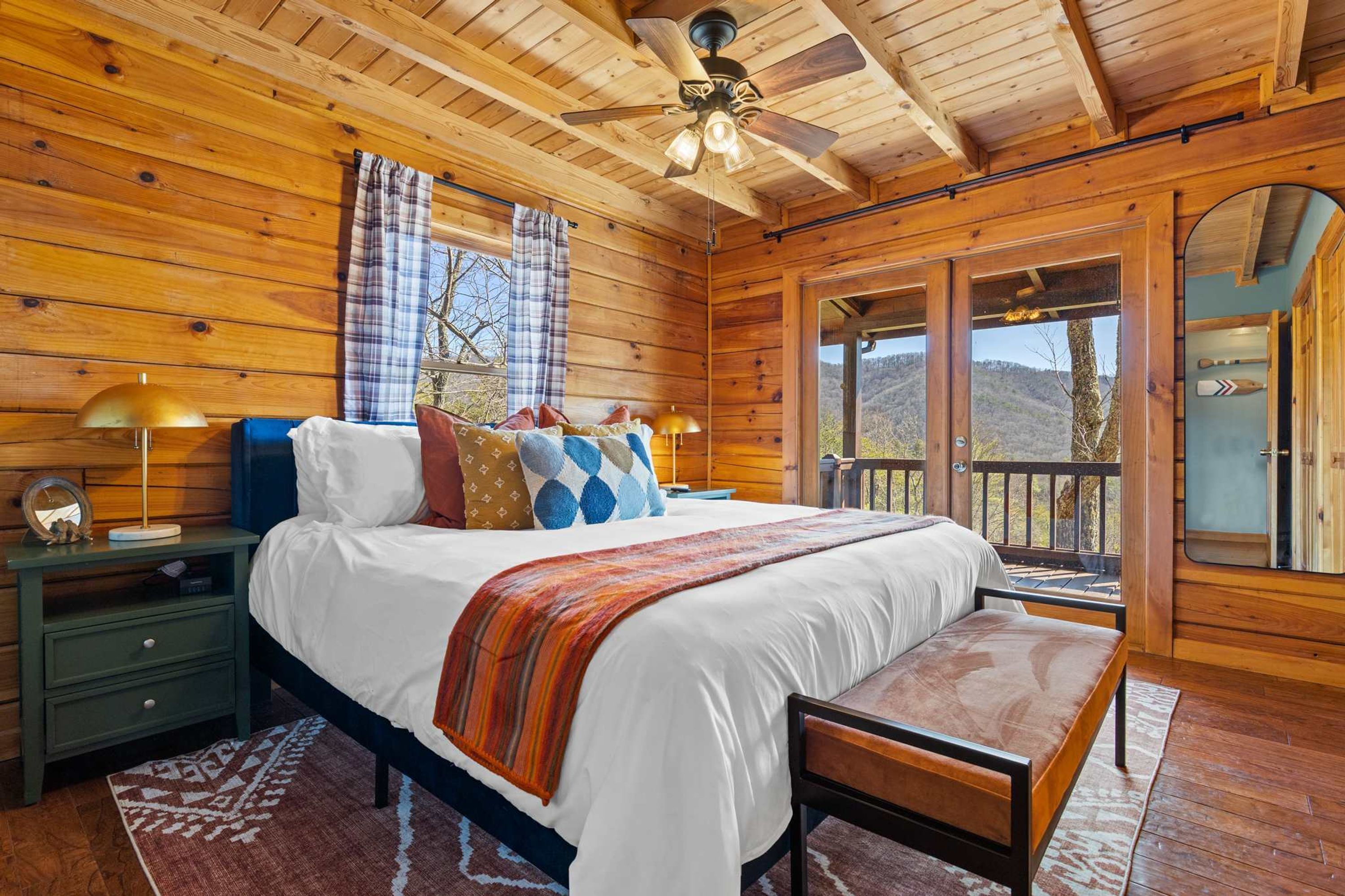 A wooden cabin bedroom with a bed, nightstands, a bench, and a view of the outdoors.