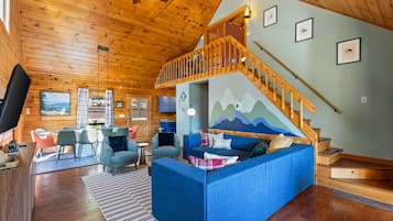 A cozy cabin interior with a wooden ceiling, a blue sofa, a dining area, and a staircase.