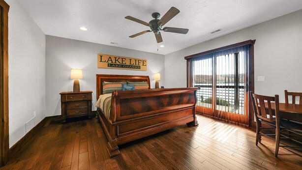 Un dormitorio con cama, mesitas de noche, ventilador de techo, un cartel que dice 'LAKE LIFE LAKE OZARKS' y vistas a través de un ventanal.