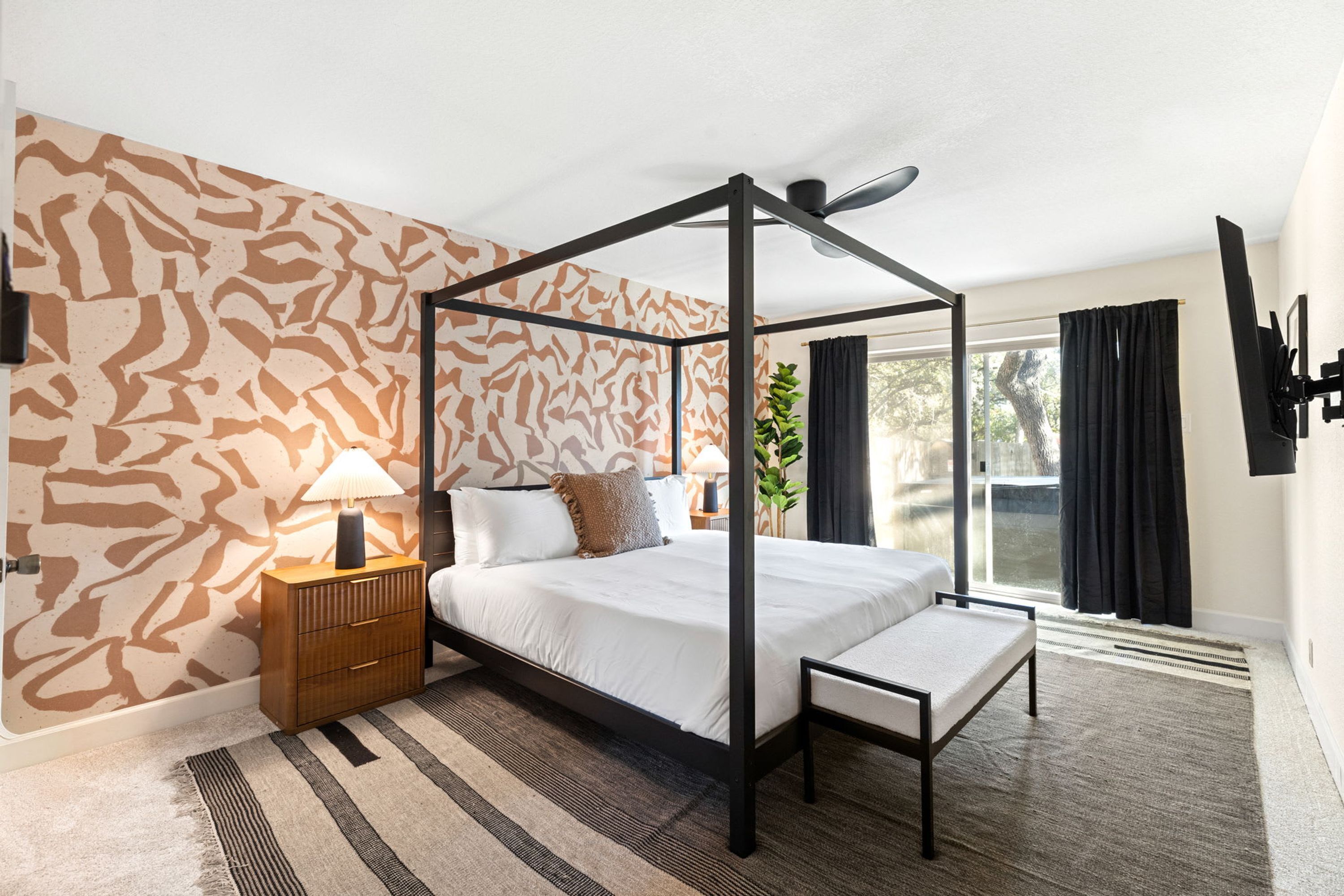 A four-poster bed with a bench, a bedside table, a lamp, and a patterned wall.