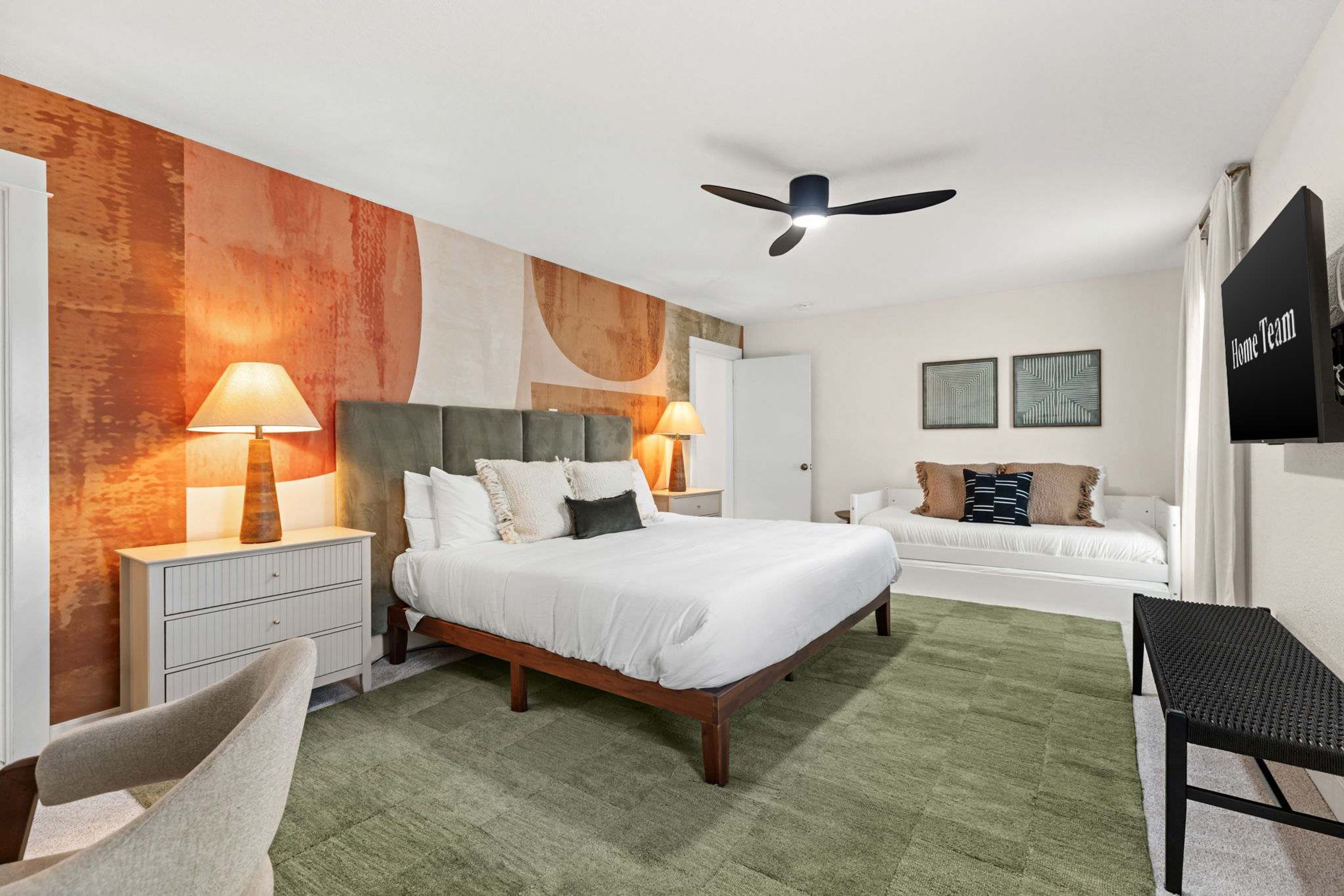A hotel room with a bed, two bedside tables with lamps, a chair, a desk, a ceiling fan, and artwork on the walls.