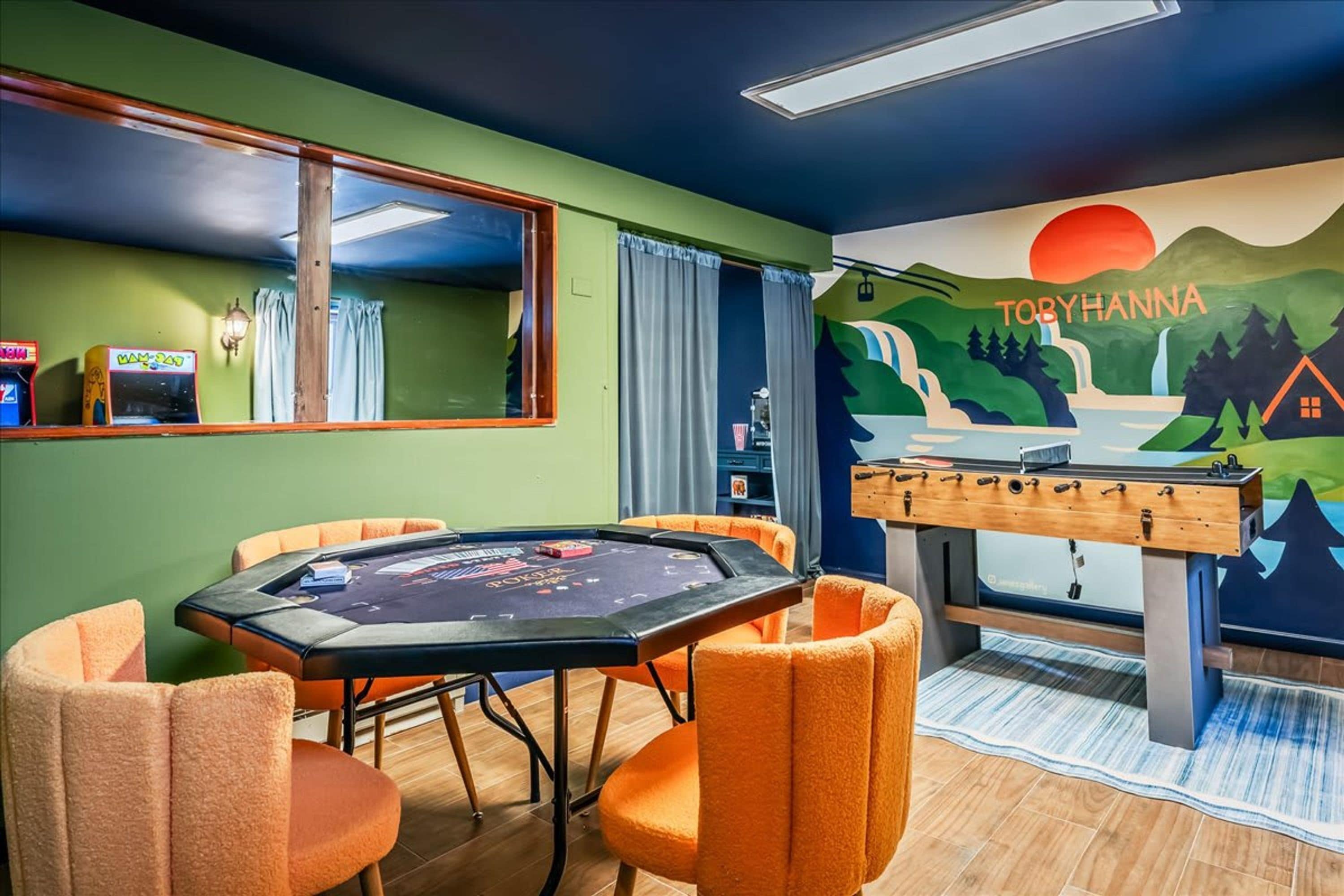 A room with a pool table, four orange chairs, a foosball table, a mural with the text 'TOBYHANNA', and a window with curtains.