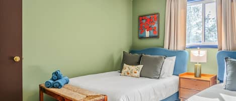 Un dormitorio con cama, mesita de noche, banca, ventana con cortinas y un cuadro en la pared.