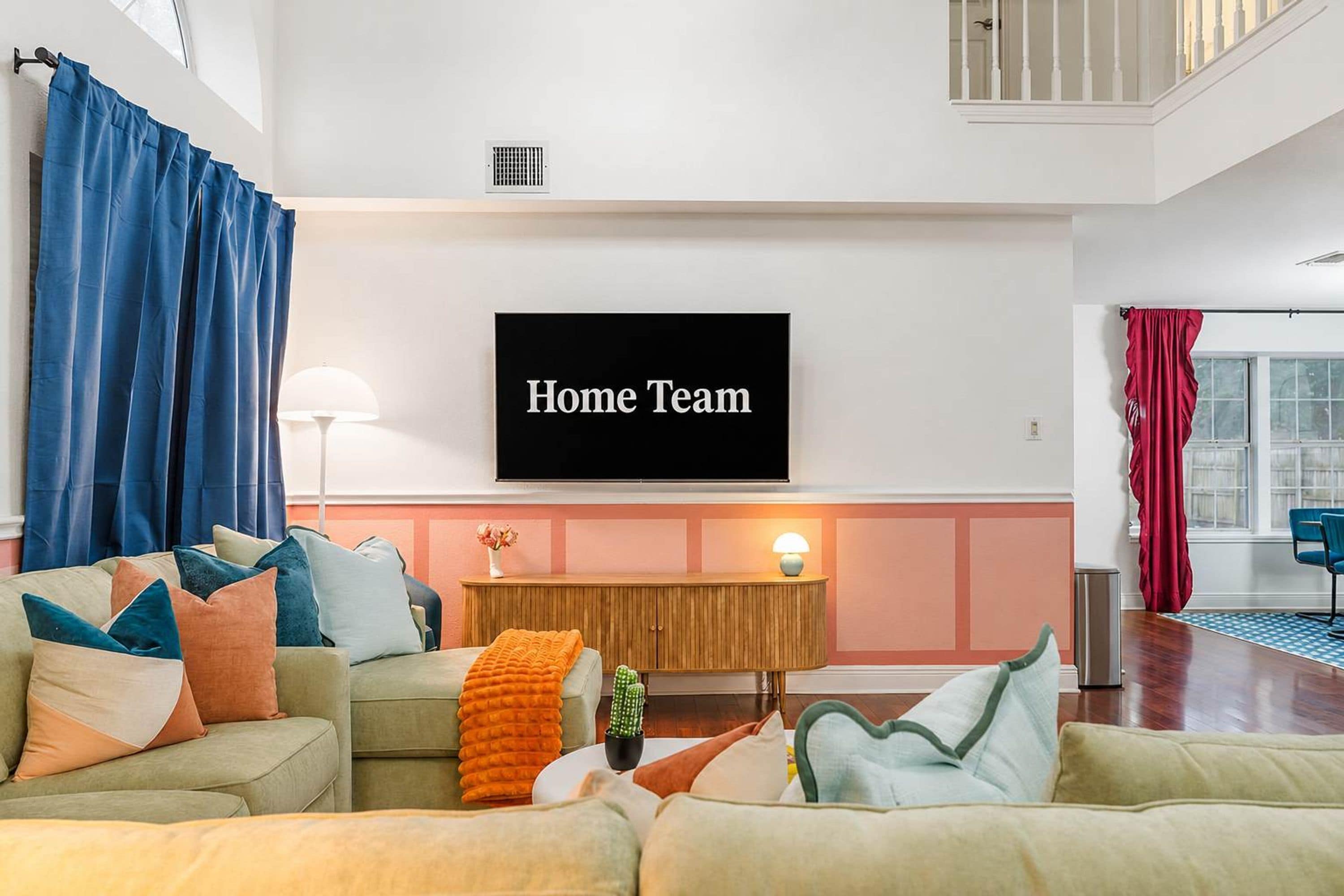 A living room with a flat-screen TV displaying 'Home Team,' a sofa set, a small table with a lamp, and a cactus plant.