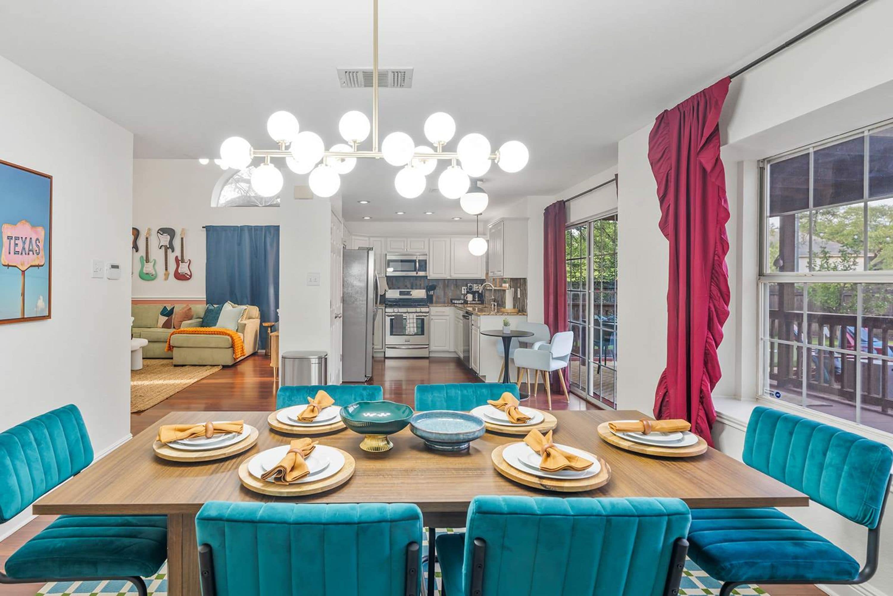 A modern dining area with a wooden table set for six, teal upholstered chairs, and a kitchen in the background.