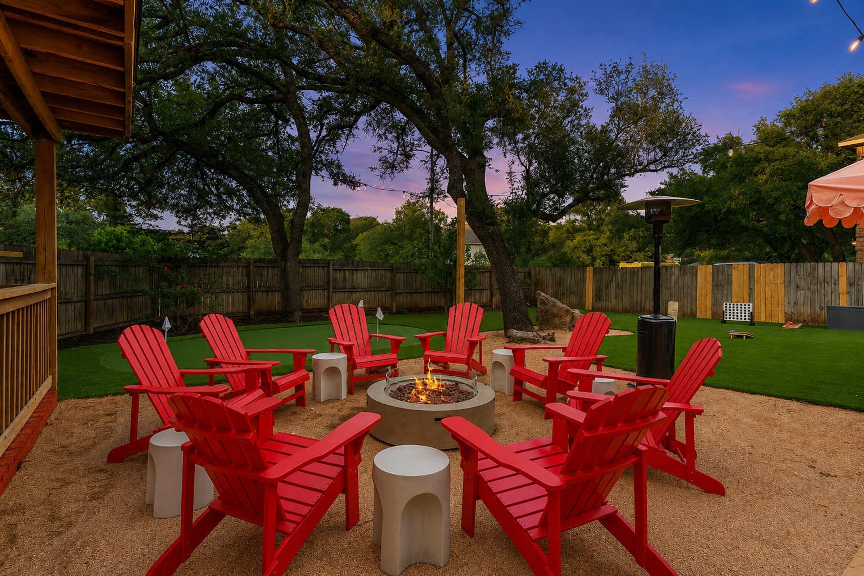 A backyard with a fire pit surrounded by red chairs and tables, a wooden deck, and a grassy area.