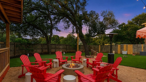 A backyard with a fire pit surrounded by red chairs and tables, a wooden deck, and a grassy area. - Austin Encore Pool Hot Tub Putt-putt (Austin)