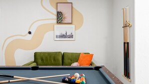 Ein Billardtisch mit Billardstöcken und Kugeln, ein grünes Sofa mit einem orangefarbenen Kissen sowie zwei gerahmte Bilder an der Wand.