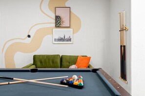 Ein Billardtisch mit Billardstöcken und Kugeln, ein grünes Sofa mit einem orangefarbenen Kissen sowie zwei gerahmte Bilder an der Wand.