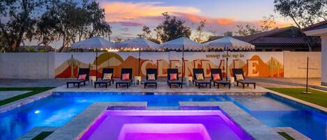 A pool area with lounge chairs and umbrellas, a wall with a mural, and a sunset sky.