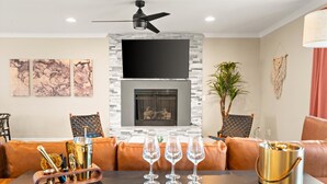 A modern living room with a fireplace, leather sofas, a dining table with wine glasses, and a ceiling fan.