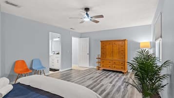 A bedroom with a bed, two chairs, a wooden dresser, a ceiling fan, and a potted plant.