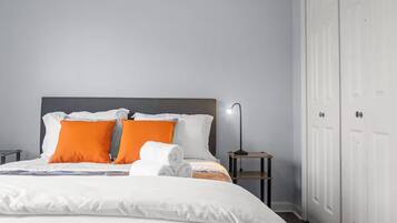 A neatly made bed with white and orange bedding, a bedside table with a lamp, and a white wardrobe.