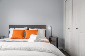 Uma cama bem arrumada com roupa de cama branca e laranja, uma mesa de cabeceira com abajur e um guarda-roupa branco.
