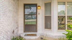 Uma casa com fachada de tijolos brancos, porta com painel de vidro, pequeno alpendre com tapete de entrada e janela com vista para o bairro.