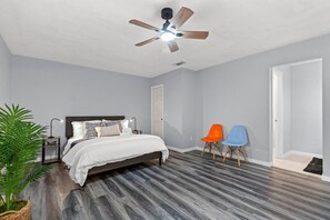Quarto com cama, ventilador de teto, duas cadeiras e um vaso de planta.
