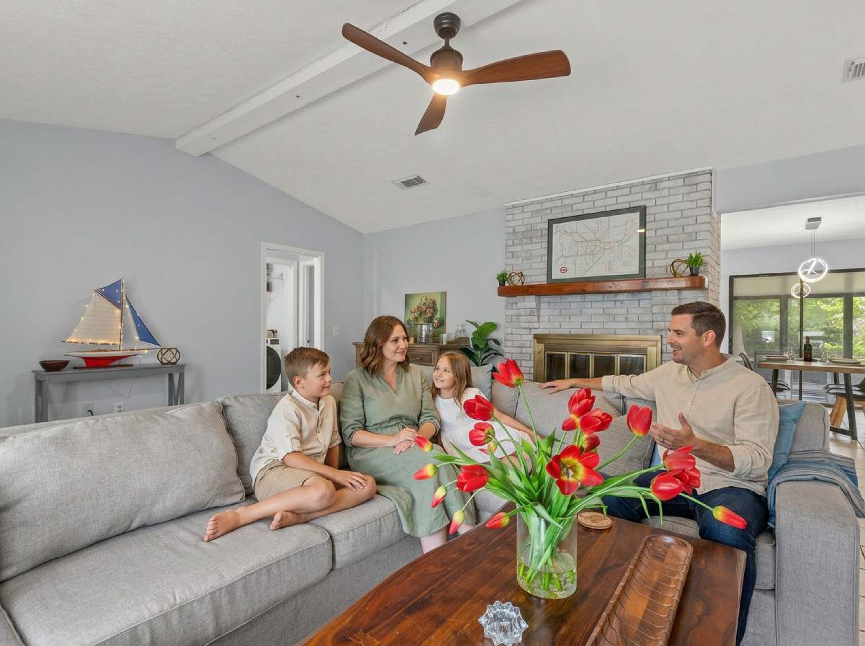 Una familia sentada en un sofá gris en una sala de estar con chimenea, un ventilador de techo y un pequeño modelo de velero sobre una mesita.