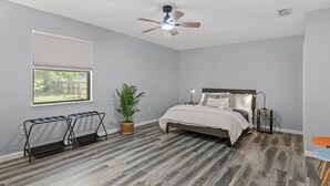 Um quarto com cama, ventilador de teto, janela com persianas, uma planta e uma mesinha de cabeceira.