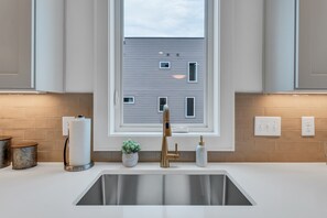 A modern kitchen with a stainless steel sink, white cabinetry, a window with a view of a building, and a potted plant on the counter. - Rooftop Nights in Nashville - 3BR - Office (Nashville)