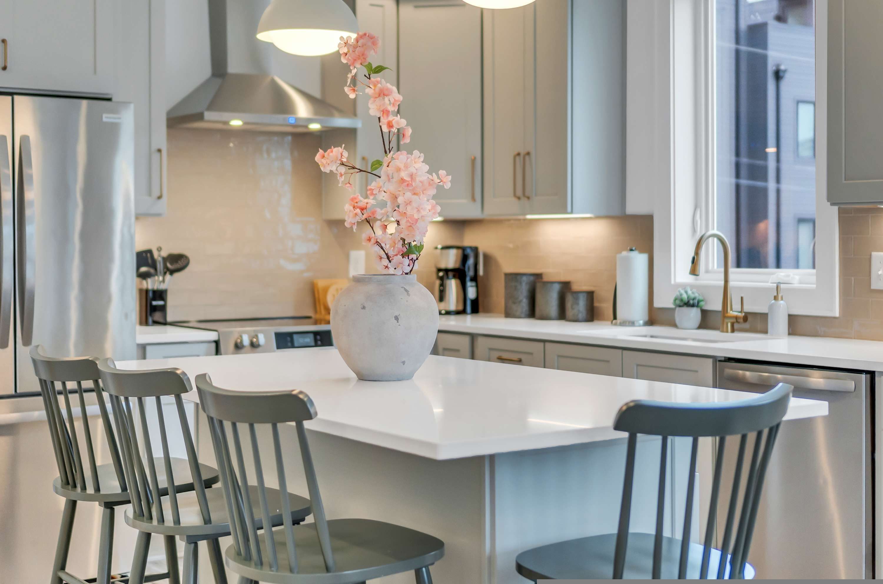 A modern kitchen with a central island, grey chairs, and a vase with pink flowers.
