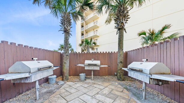 An outdoor patio area with two barbecue grills, a wooden fence, and palm trees. - Beach Front 7th Floor Views Pools Hot Tub (Orange Beach)